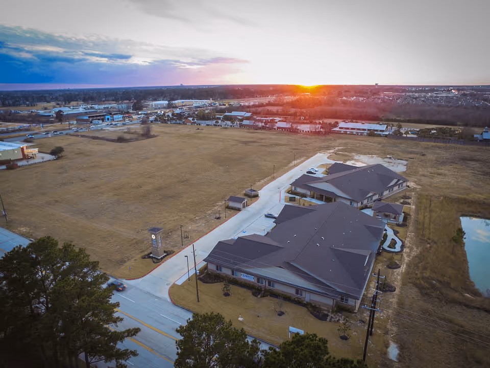 Aerial view of Countryside Senior Living of Spring facility at sunset, showing multiple buildings with dark roofs, a parking area, surrounding open grassy fields, and a road with some trees in the foreground.