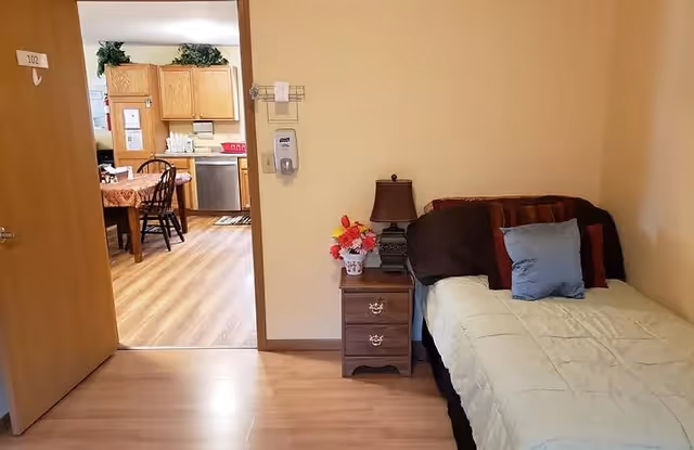 A small bedroom with a single bed covered with a light-colored blanket and several pillows. Next to the bed is a wooden nightstand with a lamp and a vase of colorful flowers. The room has light-colored walls and wooden flooring. The door to the room is open, revealing a dining area and kitchen with wooden cabinets and a dining table with chairs.