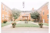 A bright central courtyard of a brick nursing home featuring a grassy lawn, paved walkways, potted flowers, trees, and a covered gazebo surrounded by three-story buildings.