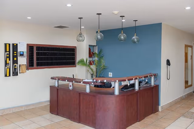 Reception desk area with a dark wood counter and a blue accent wall behind it. Four pendant lights with glass globes hang from the ceiling above the desk. A plant is placed behind the desk, and there is a wall-mounted telephone on the right wall. Mail slots and key holders are mounted on the left wall.