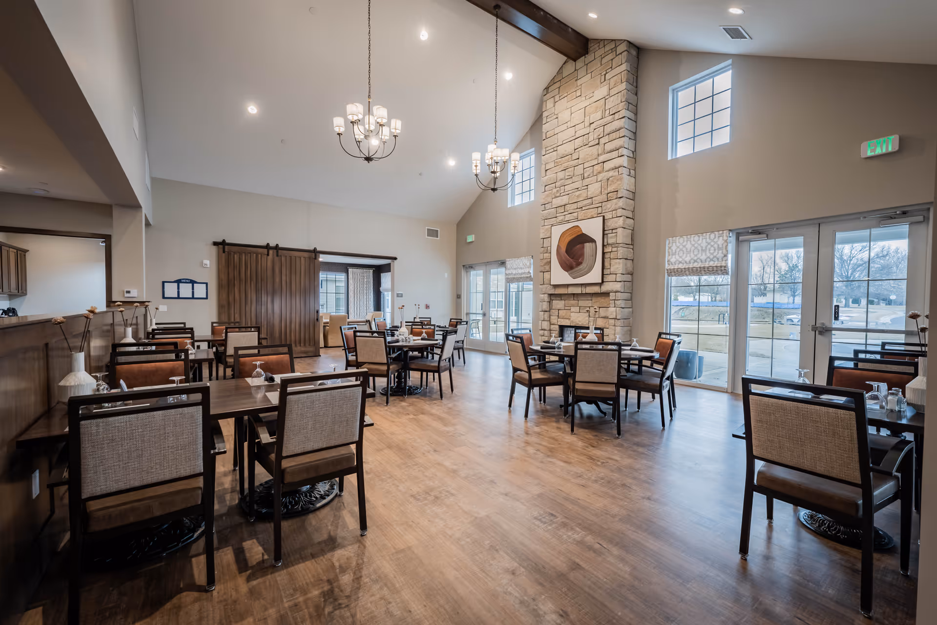 A spacious dining room in a senior living facility with multiple tables and chairs arranged neatly. The room features a high vaulted ceiling with two chandeliers, large windows letting in natural light, a stone fireplace with modern artwork above it, and wooden flooring. There is a sliding barn door leading to another room and glass doors providing access to an outdoor area.