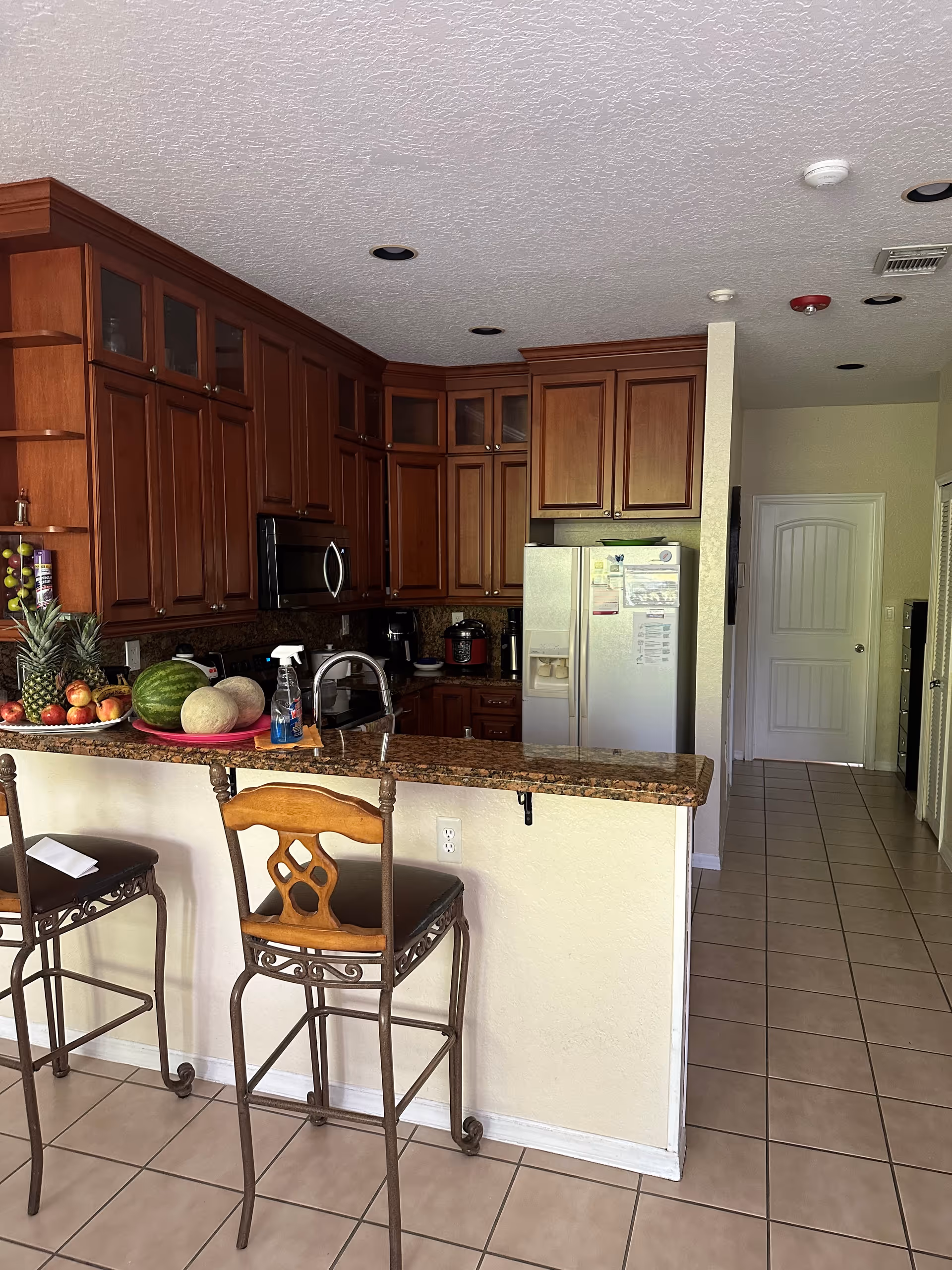 Interior view of a kitchen with wooden cabinets, a granite countertop bar with two high chairs, a refrigerator, microwave, and various fruits and kitchen items on the counter. The floor is tiled and there is a hallway leading to a white door.