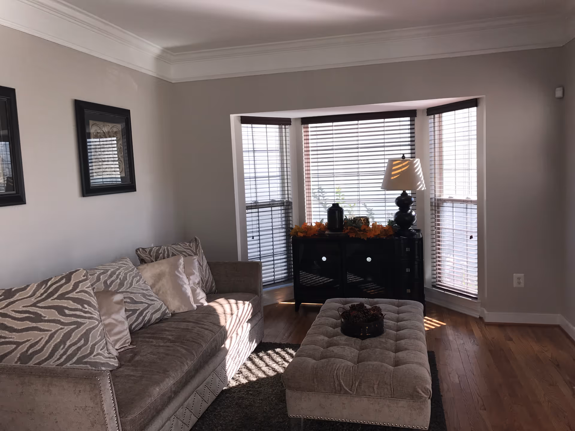A cozy living room with a beige sofa adorned with patterned and plain cushions, a tufted ottoman in front, and a black cabinet under a bay window with blinds. A table lamp and decorative items are placed on the cabinet. The room has wooden flooring and light gray walls with two framed pictures hanging.