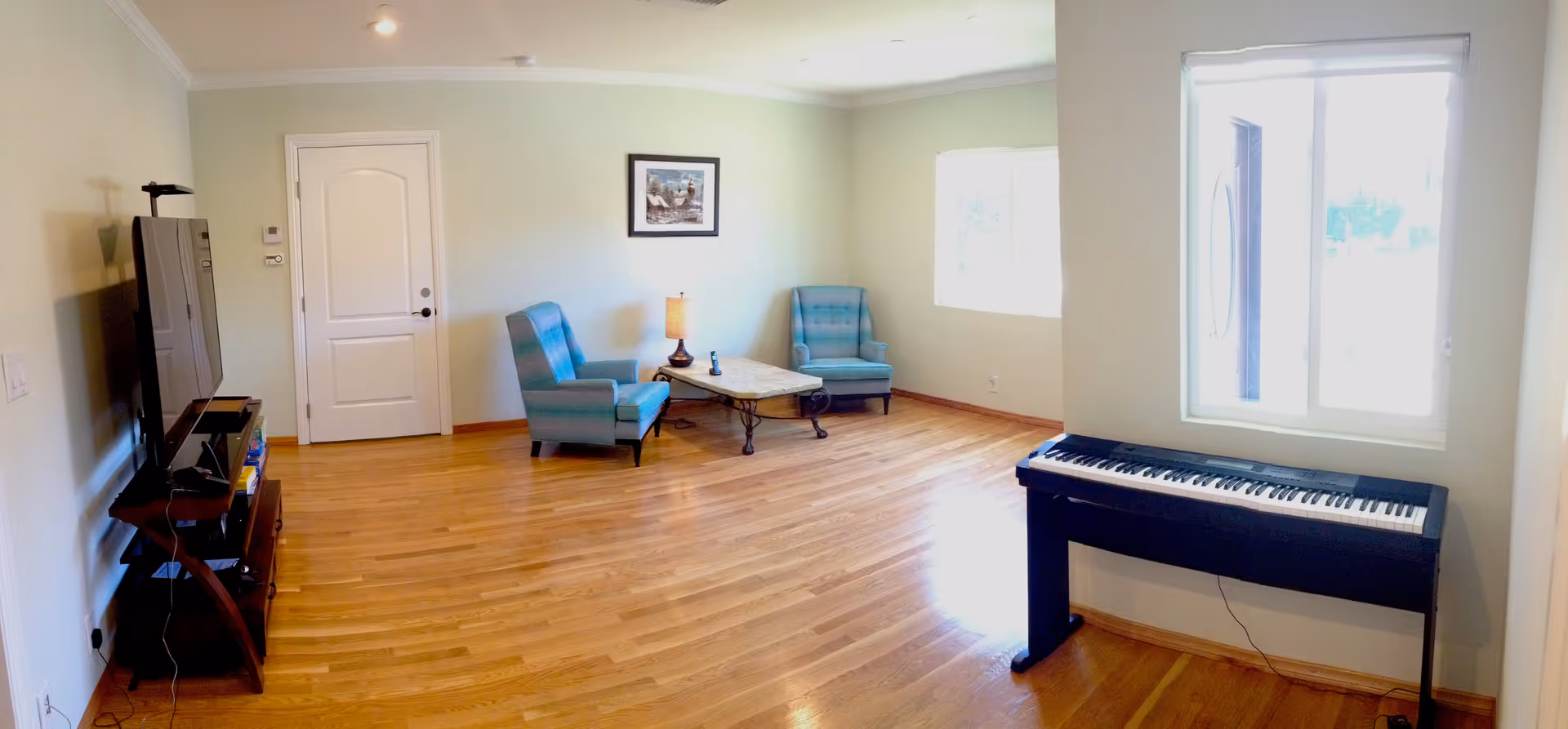 A bright living room with light green walls and wooden flooring. The room features two blue armchairs with a small table and lamp between them, a flat-screen TV mounted on a stand to the left, and a digital piano against the wall near a window on the right. A framed picture hangs on the wall above the armchairs.