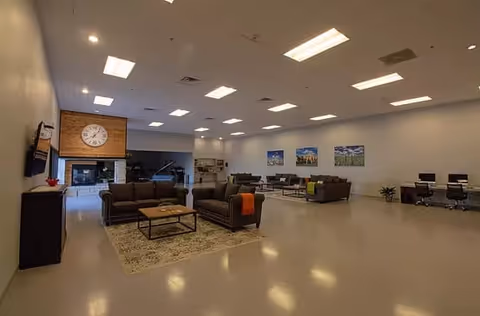 Spacious assisted living common area with multiple seating arrangements including sofas and armchairs around coffee tables, a large clock above a fireplace, a piano in the background, and a desk area with computers along the far wall. The room has bright overhead lighting and neutral-colored walls and flooring.