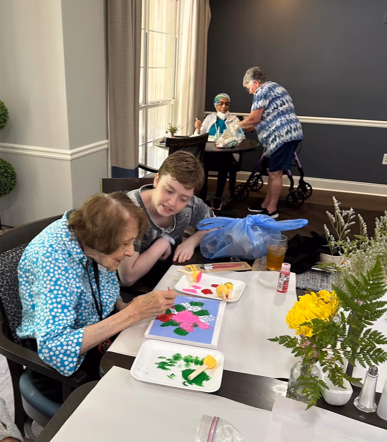 An elderly woman and a younger person sitting at a table engaged in painting a floral picture with pink and green colors. In the background, another elderly woman with a walker is standing near a seated woman at a small round table. The room has gray walls, large windows with curtains, and a vase with yellow flowers on the table.