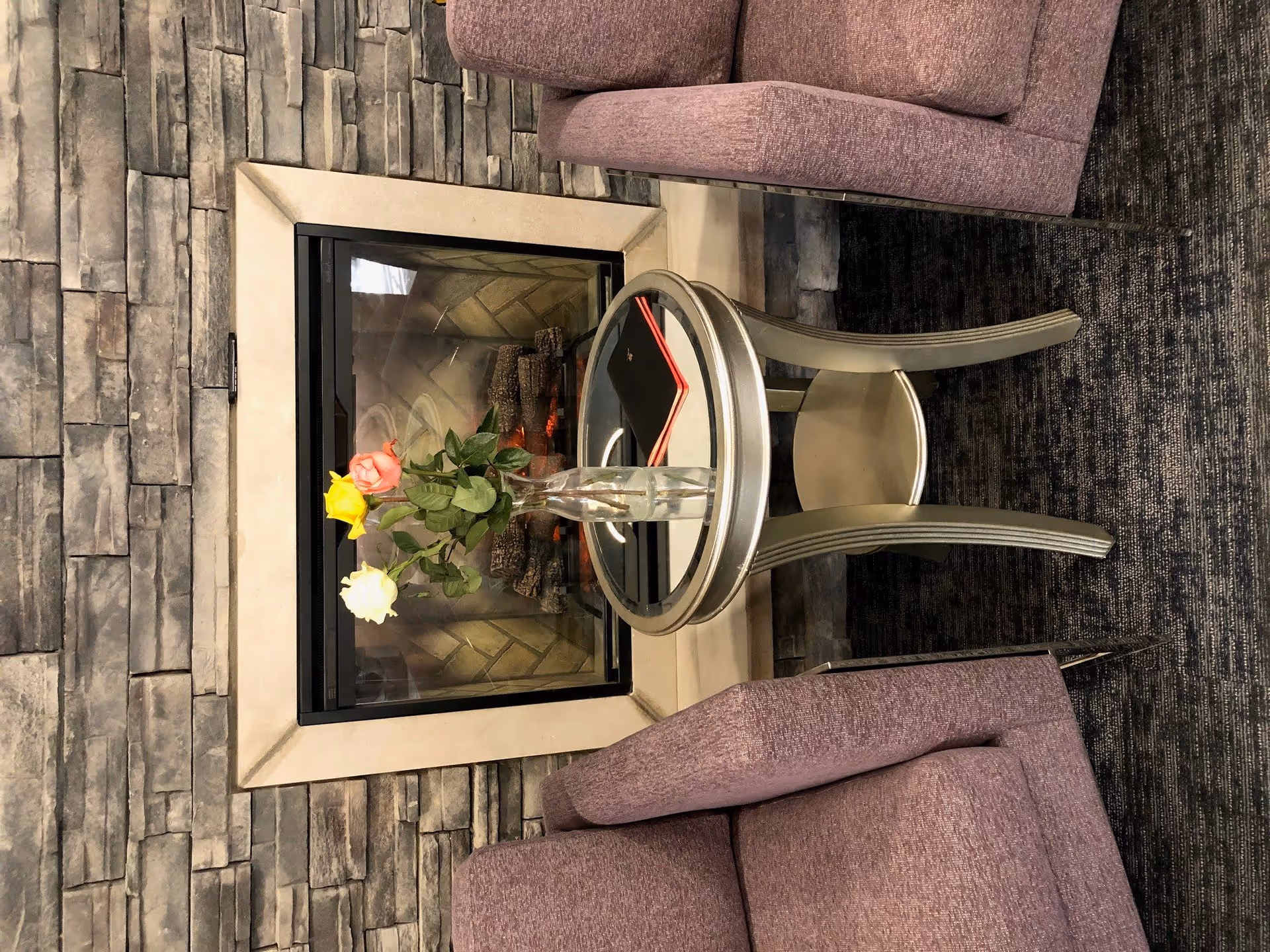 Two purple armchairs flank a round glass-topped side table holding a vase of roses in front of a stone fireplace.