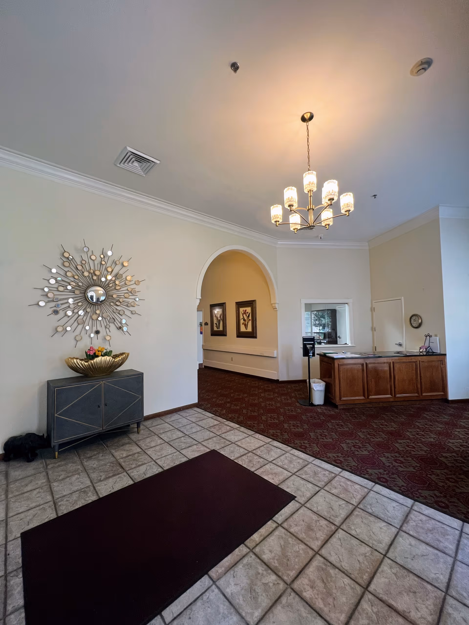 Interior view of a reception area in a senior living facility with tiled floor and a maroon carpet. There is a decorative wall mirror above a small cabinet with a gold bowl and flowers. A chandelier with multiple lights hangs from the ceiling. An archway leads to a hallway with framed artwork on the walls. A wooden reception desk is visible on the right side with a small window behind it.