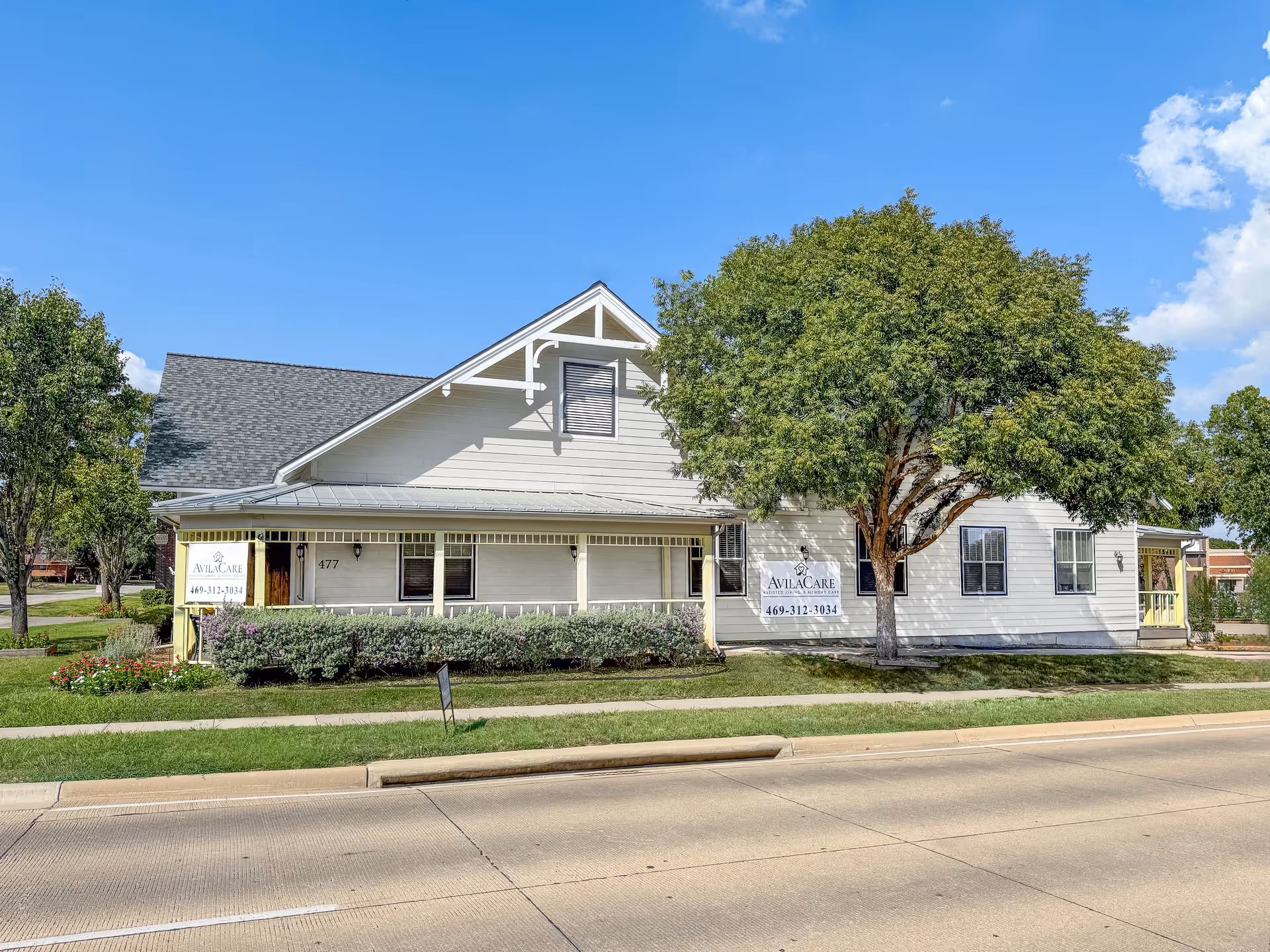 Exterior view of a white building with a gray roof and a large tree in front. The building has signs that read 'AvilaCare Memory Care of Lewisville' along with a phone number. The building is situated next to a road with a sidewalk and some landscaping including bushes and flowers.