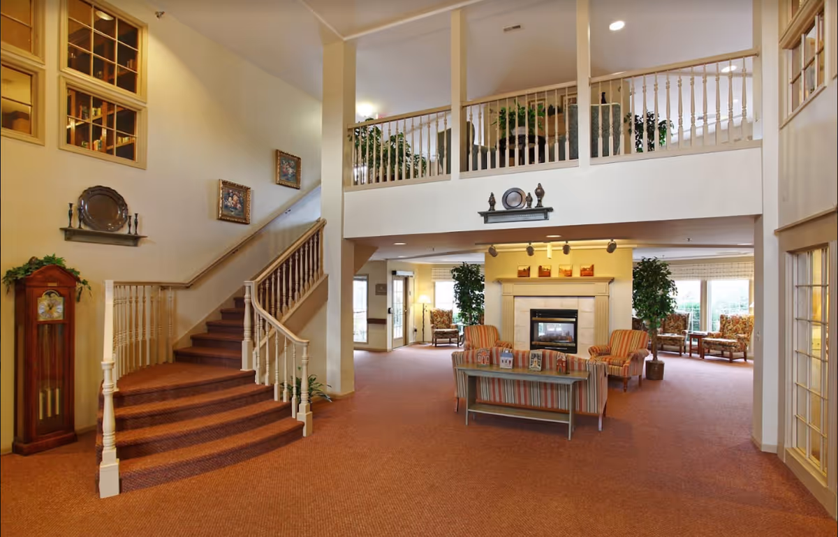 Spacious and warmly lit interior of an assisted living facility featuring a carpeted floor, a staircase with wooden railings, a fireplace surrounded by comfortable chairs and a sofa, decorative plants, and artwork on the walls. The area has a high ceiling with a balcony overlooking the space.