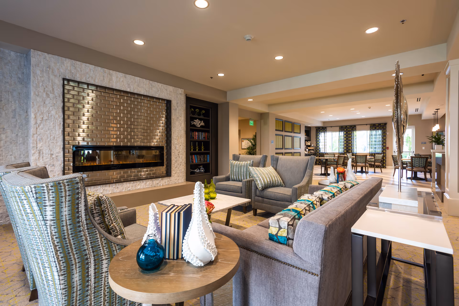 A modern senior living facility common area with comfortable seating including armchairs and a sofa, a decorative fireplace with a metallic tile surround, a round wooden table with decorative items and books, and a dining area with tables and chairs in the background. The space is well-lit with recessed ceiling lights and large windows with patterned curtains.