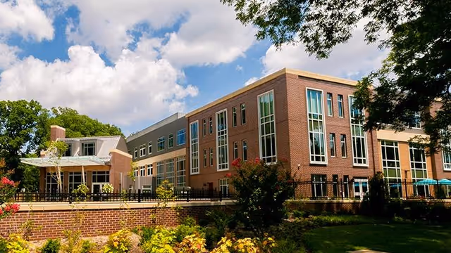 Exterior view of a modern brick and glass senior living facility building surrounded by landscaped gardens and trees under a partly cloudy sky.