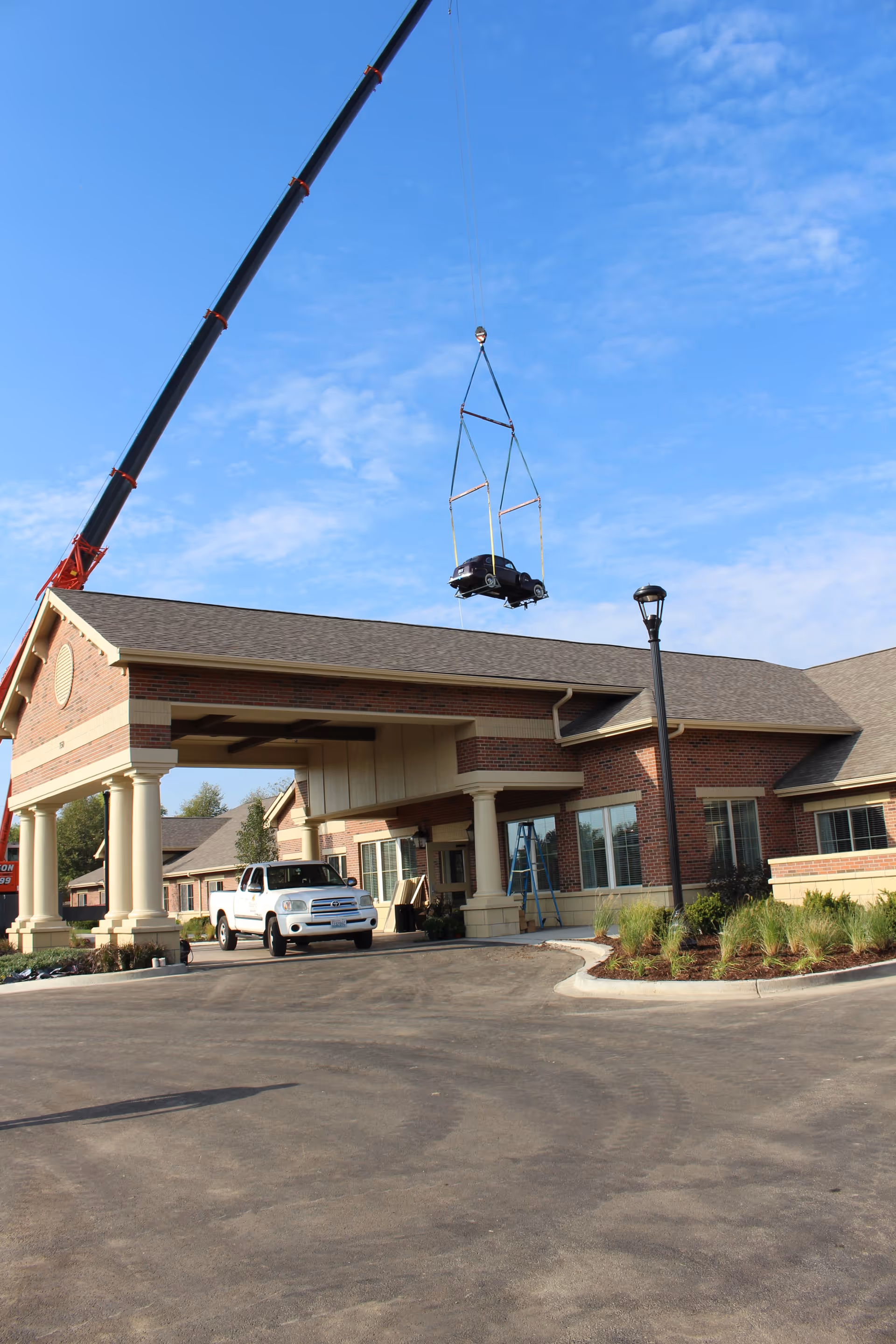 A crane is lifting a small black car above the entrance of a brick building with columns and a covered driveway. A white pickup truck is parked under the covered entrance, and there is a streetlamp and some landscaping with plants near the driveway. The sky is clear with some clouds.