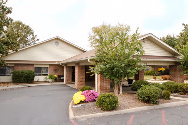 Exterior view of Easley Place facility showing a single-story building with a covered entrance supported by brick columns. There are bushes, small trees, and colorful flowers planted around the entrance area, and a paved driveway in front.