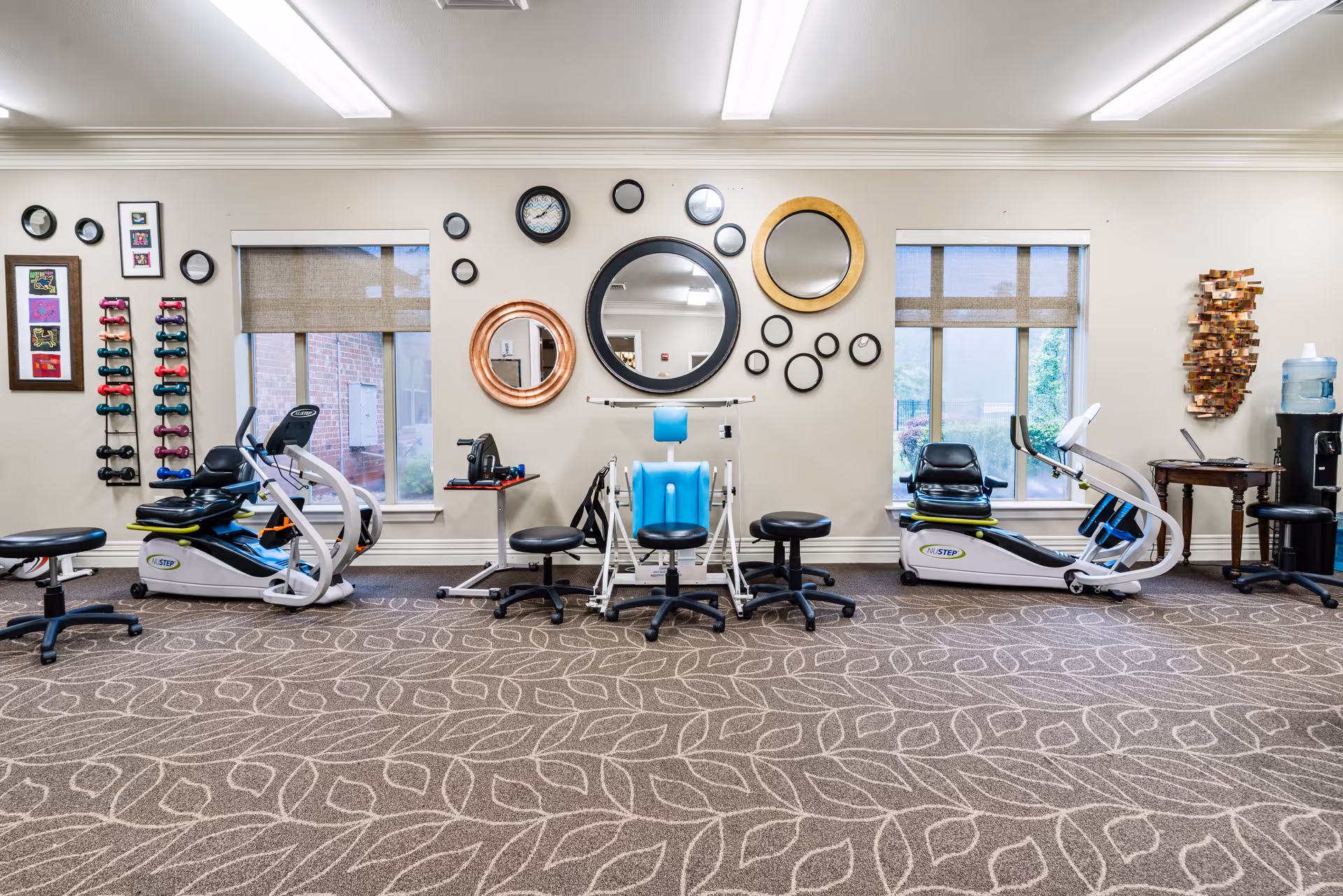 A rehabilitation exercise room with two recumbent exercise bikes, a blue therapy chair, stools, and a rack of colorful hand weights. The wall behind features multiple decorative round mirrors and two windows with beige blinds. There is also a water dispenser and a small table with a laptop on the right side.