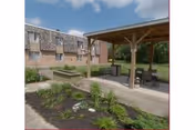 Outdoor patio area with a wooden pergola covering a seating area that includes tables and chairs. The patio is adjacent to a multi-story brick building with several windows. There are landscaped garden beds with green plants and mulch surrounding the patio. The sky is partly cloudy.