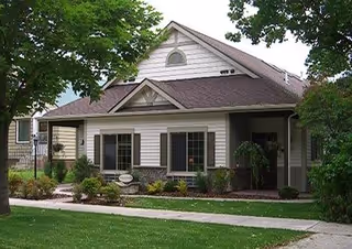 Exterior view of a single-story residential building with beige siding and a brown roof, surrounded by green trees and shrubs, with a concrete walkway leading to the entrance.