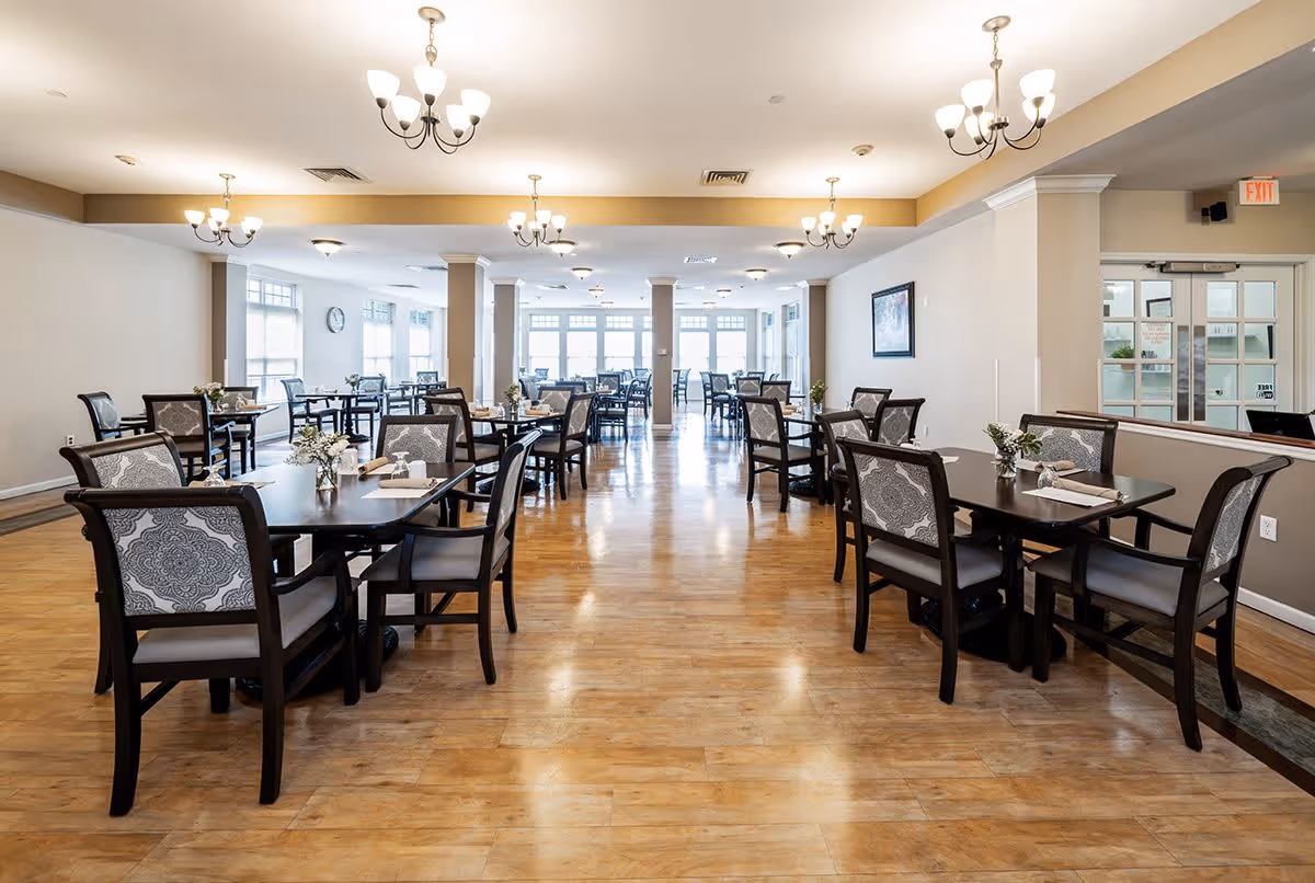 A spacious, well-lit dining room with multiple dark wood tables and chairs arranged neatly. Each table has a small floral centerpiece and napkins. The room features wooden flooring, beige walls, several chandeliers, large windows allowing natural light, and a clock on the wall.