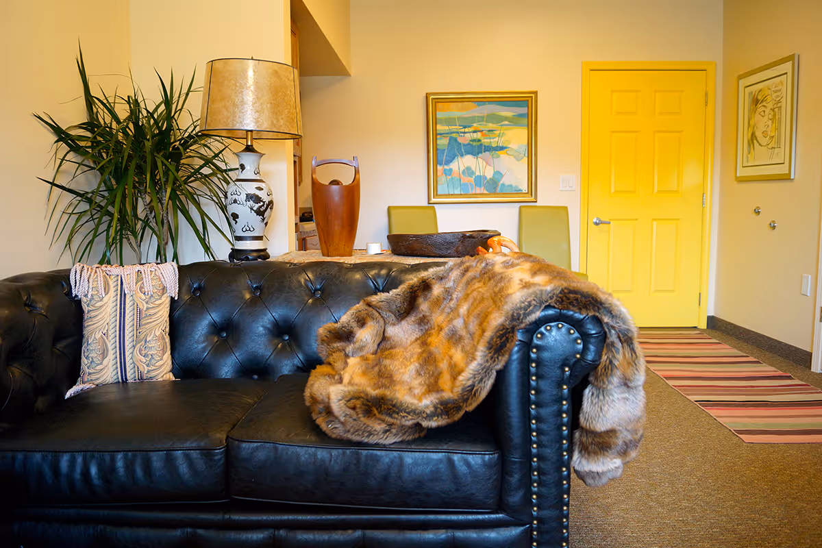A cozy living room area featuring a black leather tufted sofa with a decorative pillow and a brown fur throw draped over one arm. Behind the sofa is a table with a large ceramic lamp, a wooden decorative piece, and two green chairs. The walls are painted a warm yellow and adorned with two framed artworks. A yellow door and a striped rug are visible in the background.
