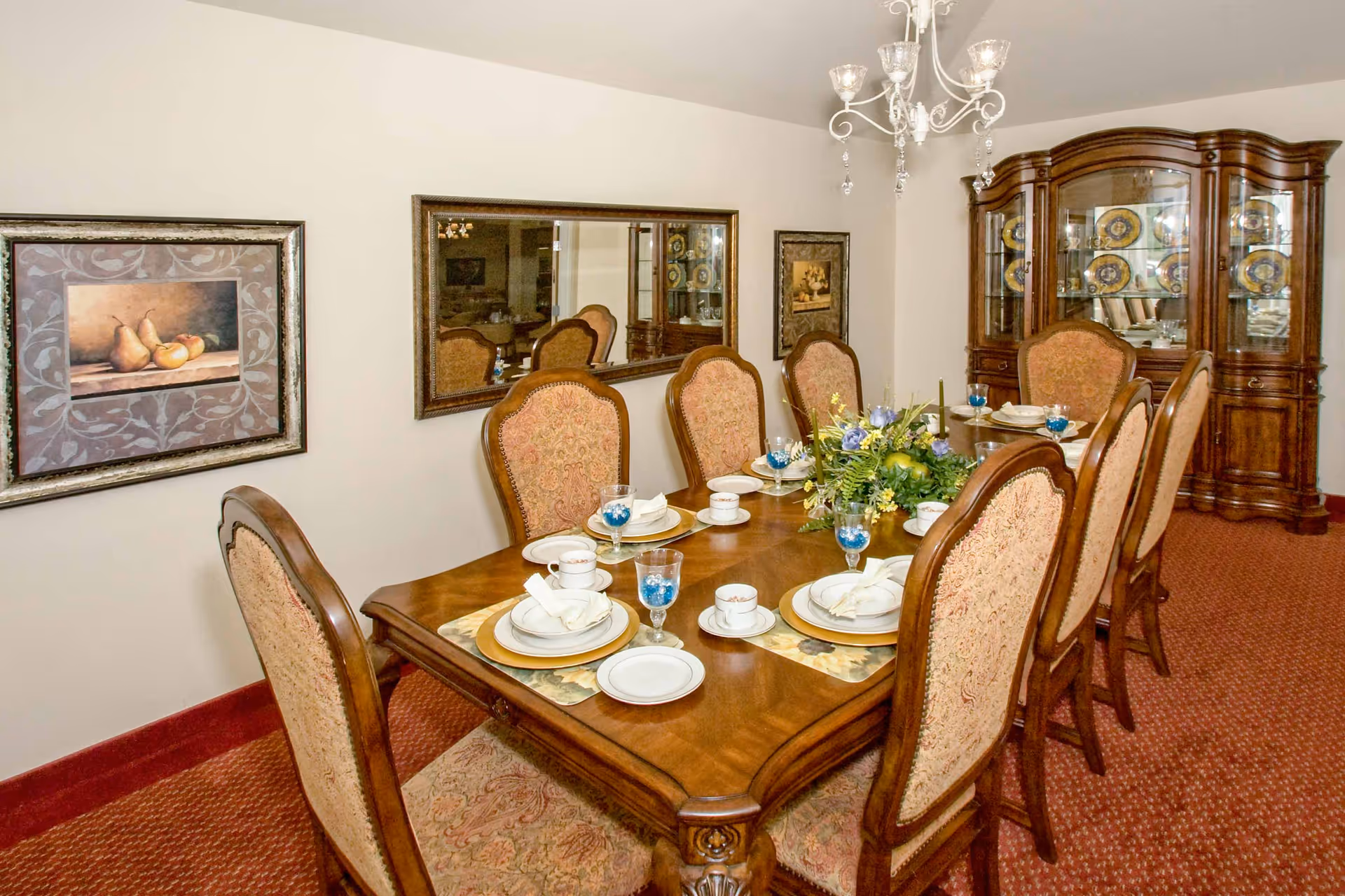 Formal dining room with an ornate wooden table set for dinner, upholstered chairs, a chandelier, mirror on the wall, and a china cabinet.