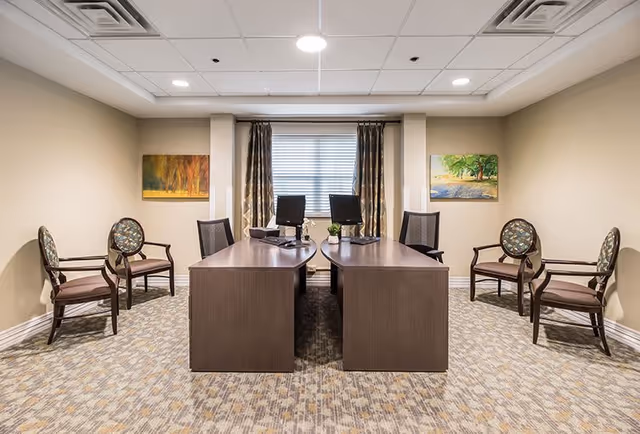 A well-lit interior office/reception area with two desks facing each other, computers, several chairs, a window with curtains, and framed artwork on the walls.