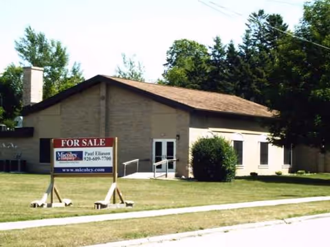 Single-story beige building with a 'For Sale' sign on a grassy front yard.