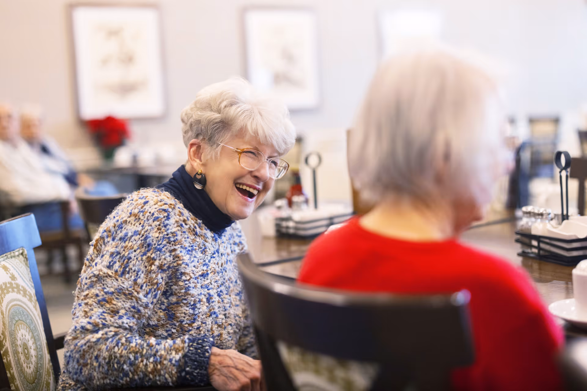 Two elderly women laugh and converse while seated at a dining table in a senior living facility.