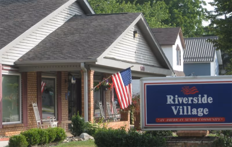 Exterior view of Riverside Village senior living facility showing a brick building with a porch, rocking chairs, an American flag, and a sign in front that reads 'Riverside Village An American Senior Community'.