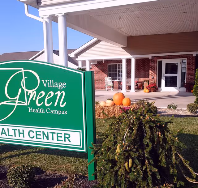 Entrance of Village Green Health Campus showing a green sign with the facility name, a covered porch with white columns, rocking chairs, and fall decorations including pumpkins and hay bales.