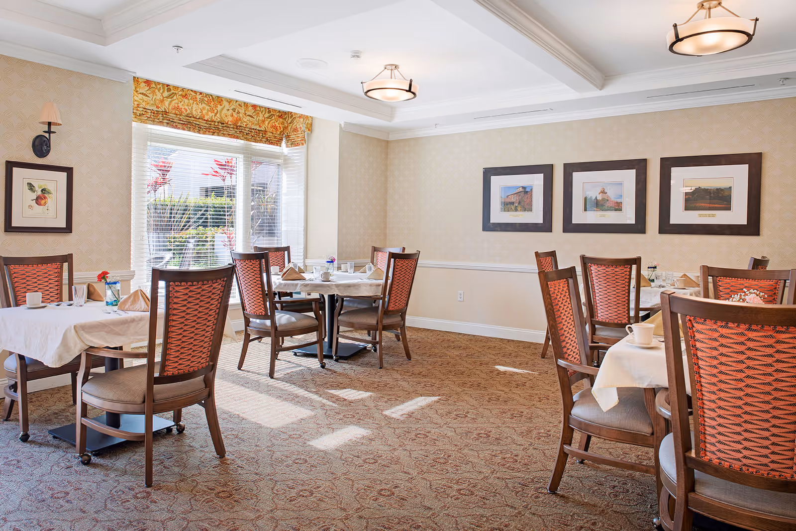 A bright dining room with several tables covered with white tablecloths and set with cups, glasses, and napkins. The chairs have wooden frames with orange patterned backs. The room has beige patterned wallpaper, a carpeted floor, framed pictures on the walls, and a large window with blinds and a floral valance letting in natural light.