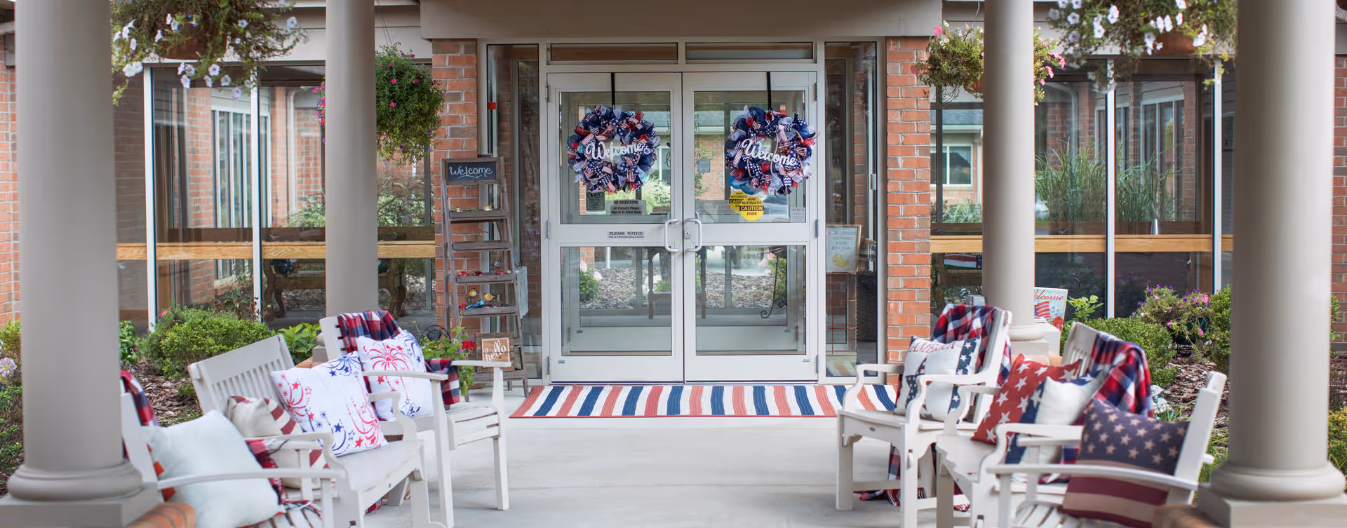 Covered outdoor seating area with white chairs decorated with patriotic red, white, and blue cushions and blankets, leading to glass double doors adorned with matching welcome wreaths. The area is framed by brick columns and hanging flower baskets.
