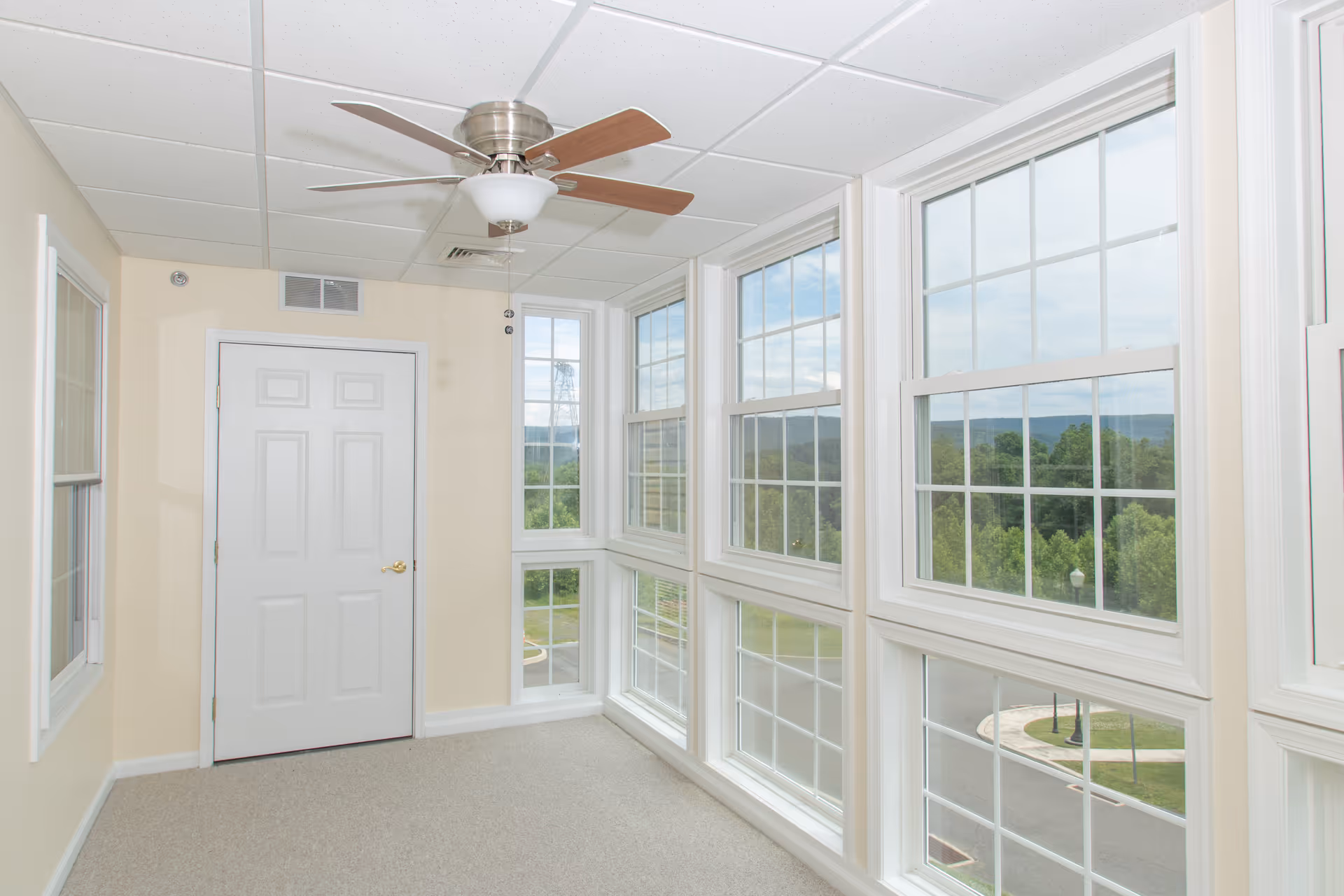 Bright interior sunroom with large grid windows along one wall, a ceiling fan, and a closed white door.