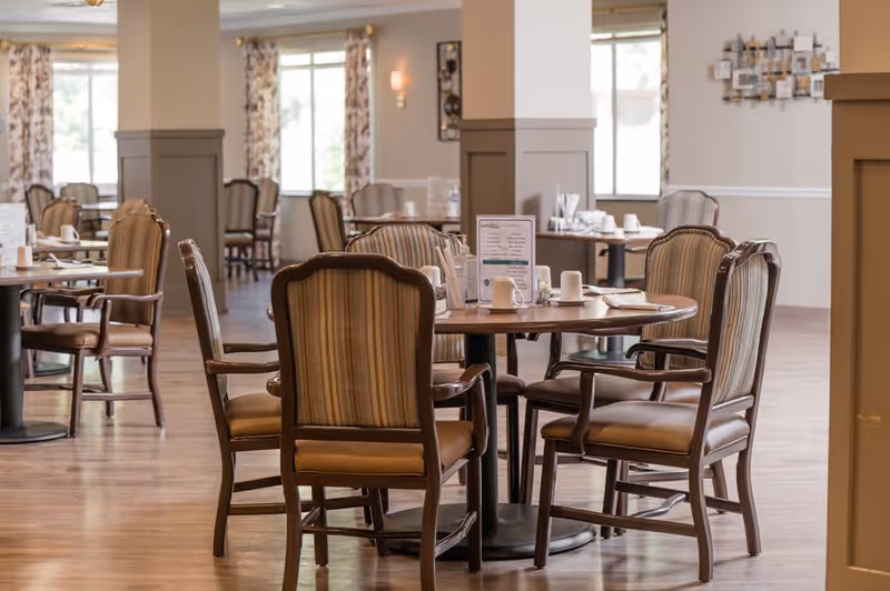 A spacious dining room with round wooden tables and cushioned chairs with striped upholstery. The tables are set with menus, cups, and napkins. Large windows with patterned curtains allow natural light to fill the room. The walls are painted in neutral tones with decorative wall art.