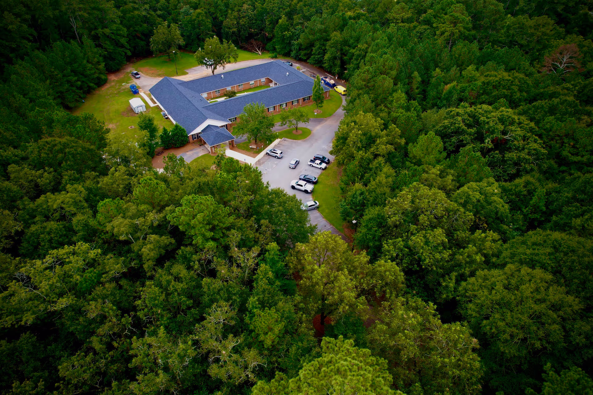 Aerial view of a single-story building with a dark roof surrounded by dense green trees. The building has a U-shaped layout with a parking lot in front containing several cars. The area is lush and forested, providing a serene and secluded environment.