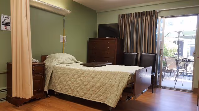 A nursing center bedroom with a single bed covered in a beige quilt, a wooden nightstand with a telephone, a tall dresser with a TV on top, two chairs, and a sliding glass door leading to an outdoor patio with a table and umbrella.