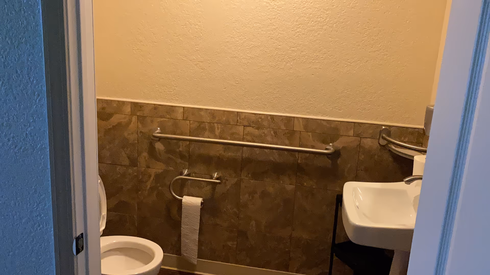 Small bathroom with beige walls and brown tiled lower half. There is a white toilet with the lid up on the left side, a metal grab bar above the toilet paper holder, and a white sink with a faucet on the right side. The bathroom has a simple and clean design.