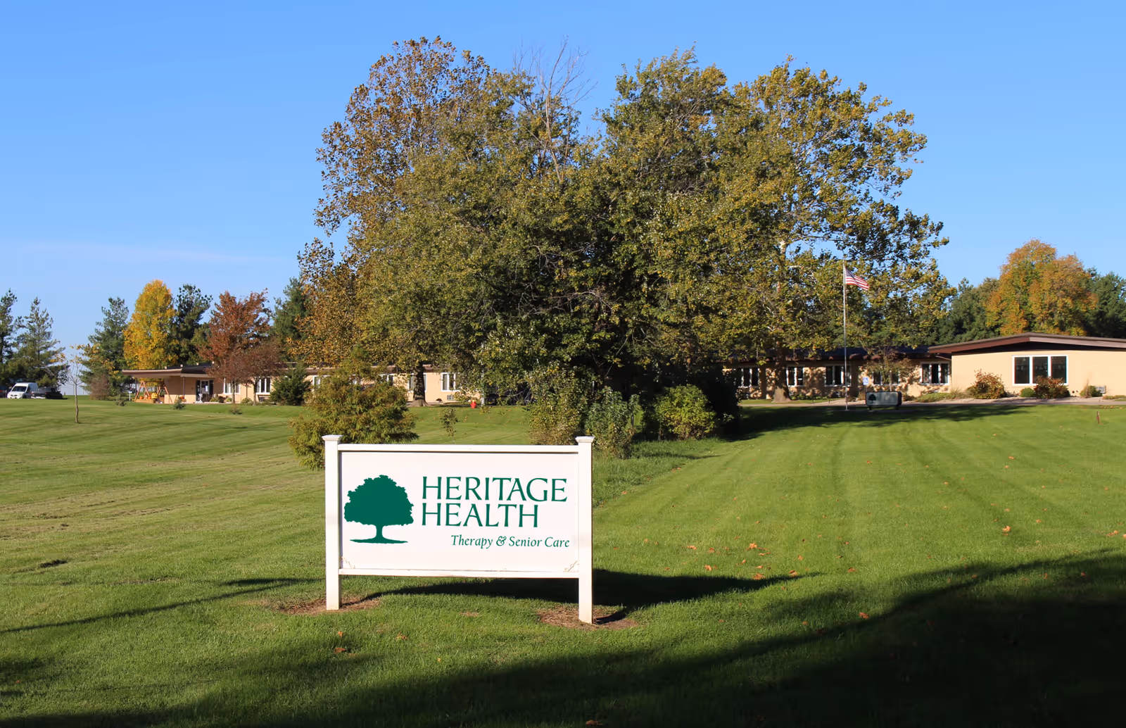 A large green lawn with a white sign in the foreground that reads 'HERITAGE HEALTH Therapy & Senior Care'. In the background, there is a single-story building partially obscured by trees, with an American flag on a flagpole near the building. The sky is clear and blue.
