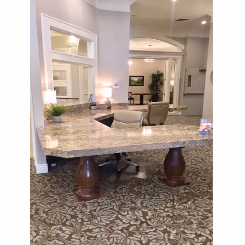 Reception desk area with a large marble countertop supported by wooden pedestals, two office chairs behind the desk, a small potted plant, and a lamp. The background shows a hallway leading to another room with a table, chairs, a plant, and a framed picture on the wall.