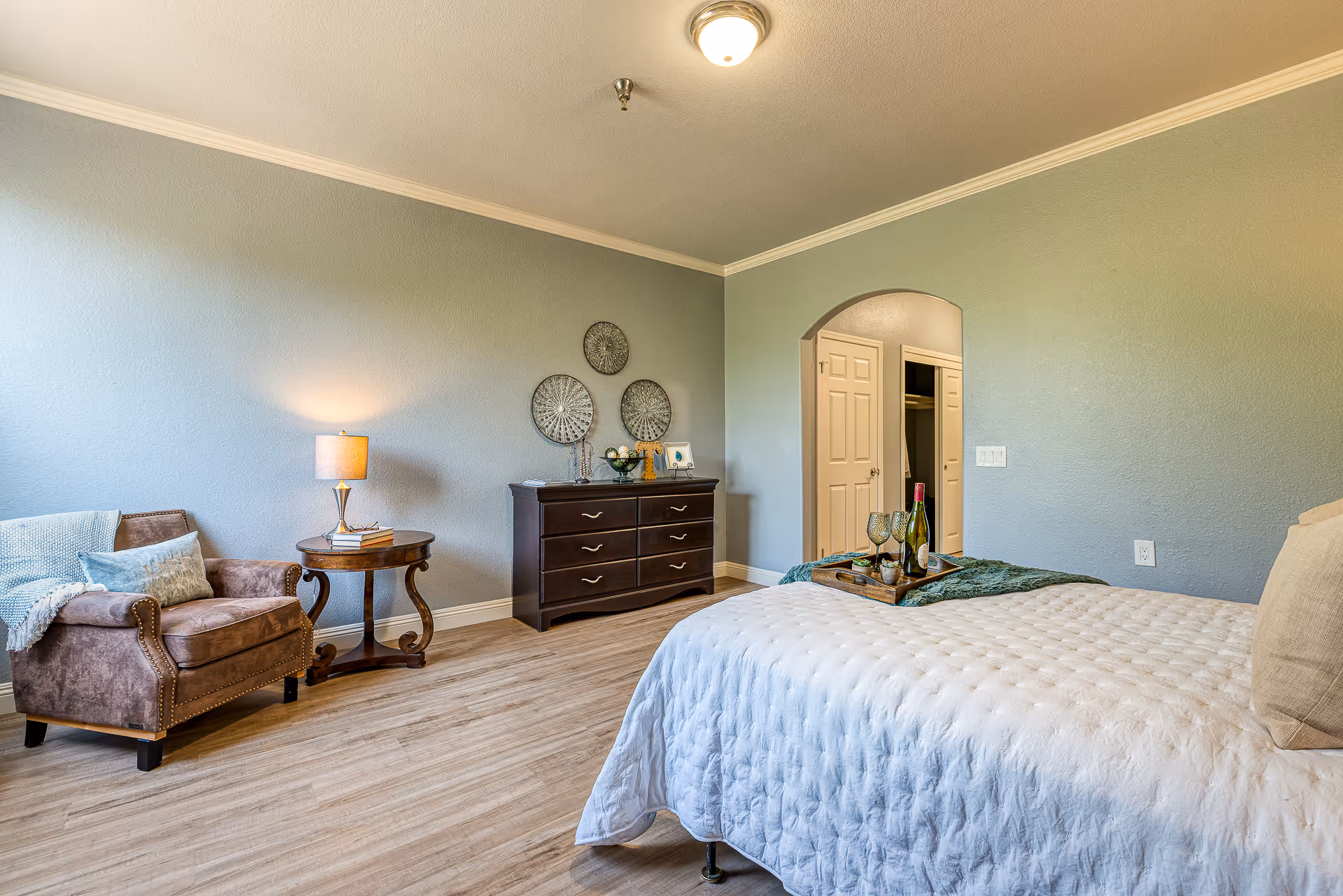 Spacious furnished bedroom with a bed in the foreground, an armchair and side table to the left, and a dresser against a blue-gray wall with an open doorway to a closet.