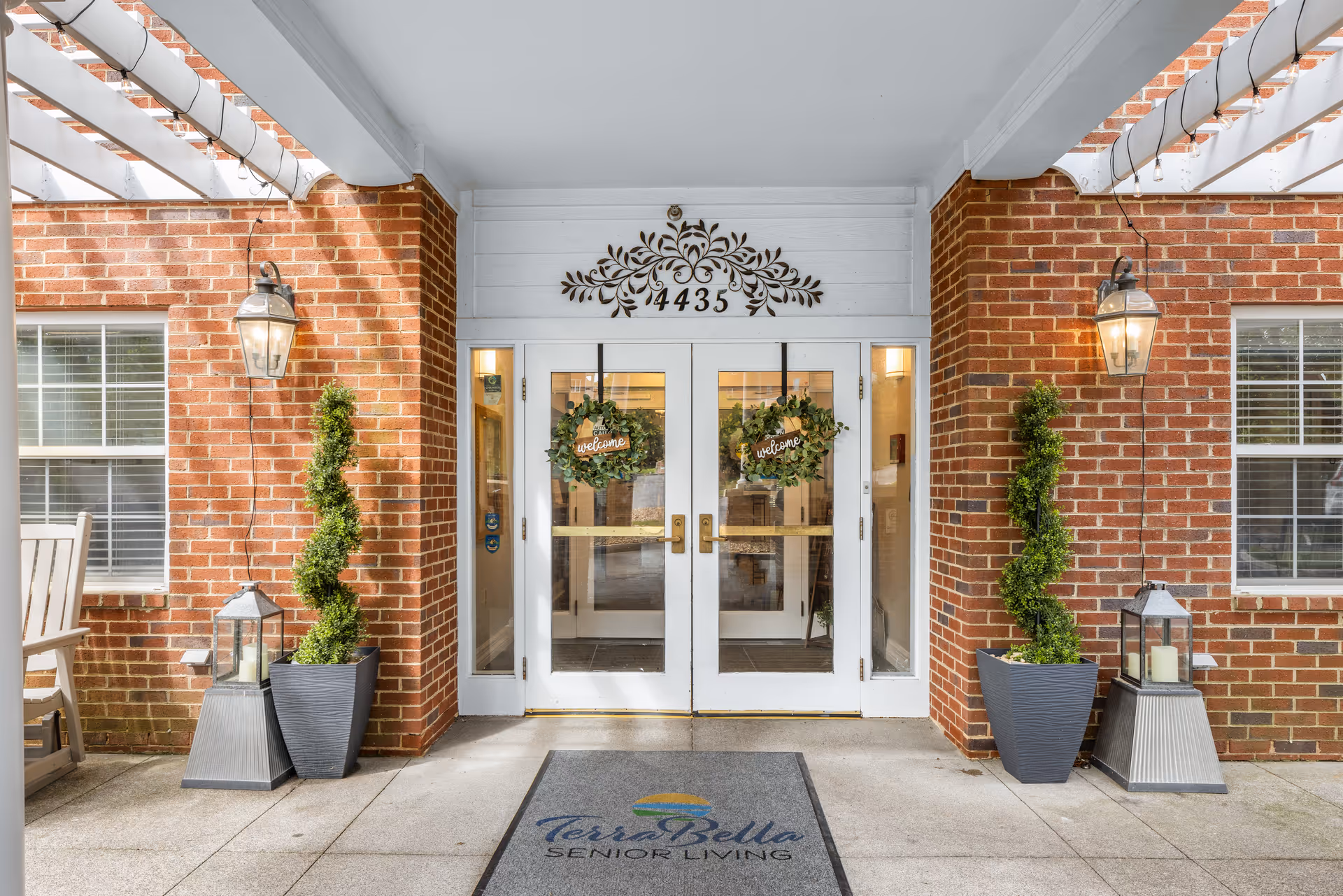 Front entrance of a senior living building with double glass doors flanked by brick walls, potted spiral topiaries, lanterns, and a welcome mat.