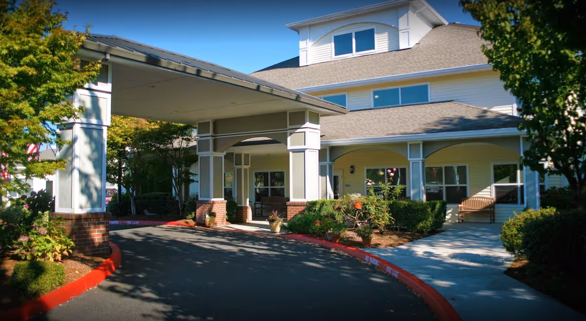 Exterior view of Prestige Senior Living Bridgewood showing the entrance with a covered driveway, surrounded by trees, bushes, and flower pots. The building has light-colored siding, multiple windows, and a bench near the entrance.