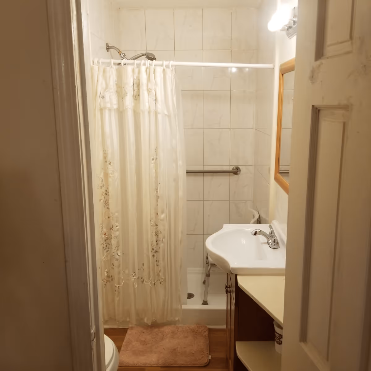 Small bathroom with a shower area enclosed by a white floral shower curtain, a white sink with a faucet, a mirror with a wooden frame, a grab bar on the tiled wall, and a pink bath mat on the floor.