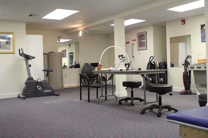 An interior therapy/exercise room with an exercise bike, tables and rolling stools, chairs and various mobility and cleaning equipment.