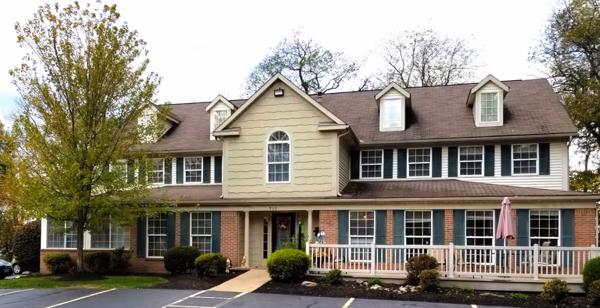 Front exterior view of a two-story assisted living facility building with beige siding, brick accents, multiple windows with green shutters, a small porch with white railing, and a tree on the left side. The parking lot is visible in the foreground.