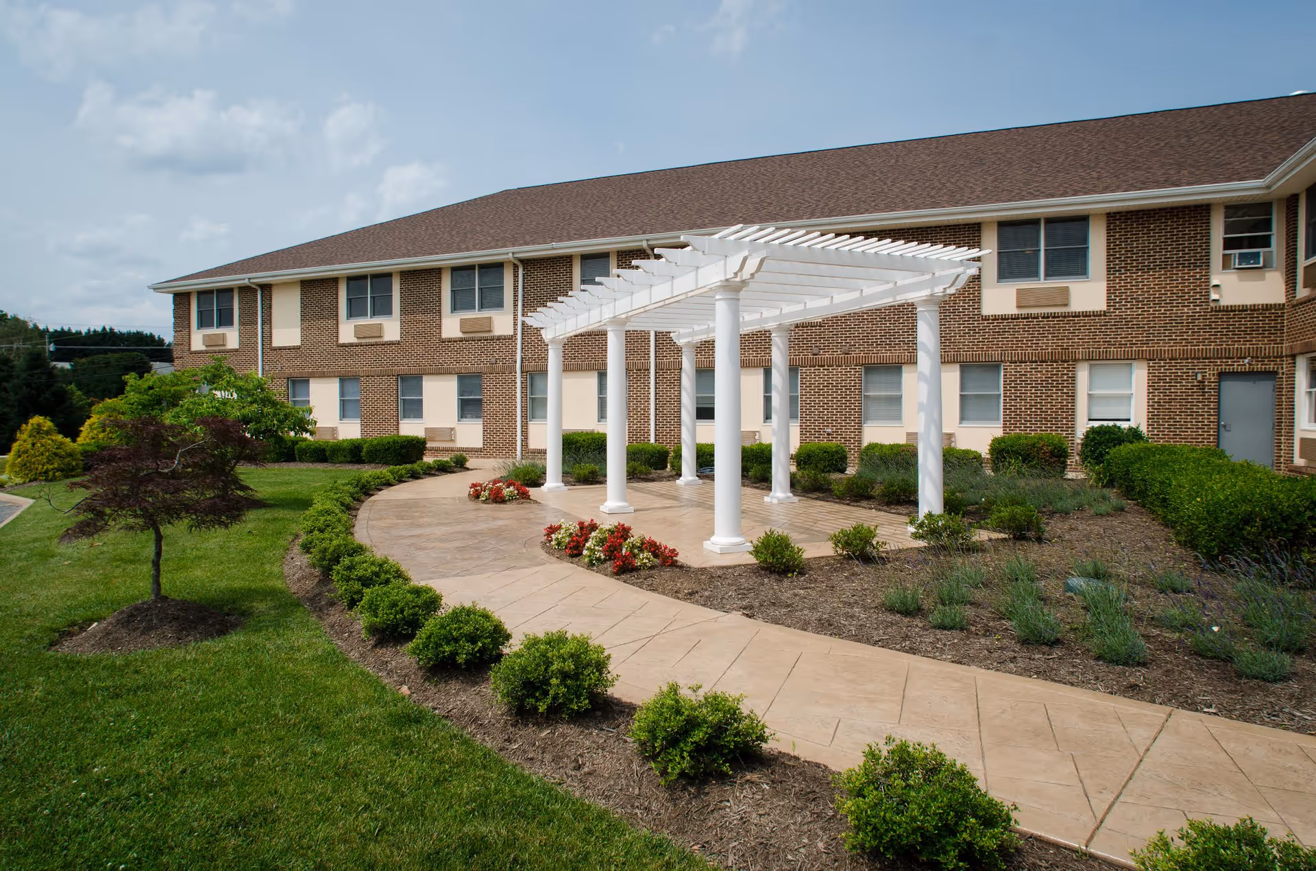 Outdoor garden area at Commonwealth Senior Living At Front Royal featuring a paved walkway, a white pergola with columns, neatly trimmed bushes, flower beds with red and white flowers, and a two-story brick building in the background under a partly cloudy sky.