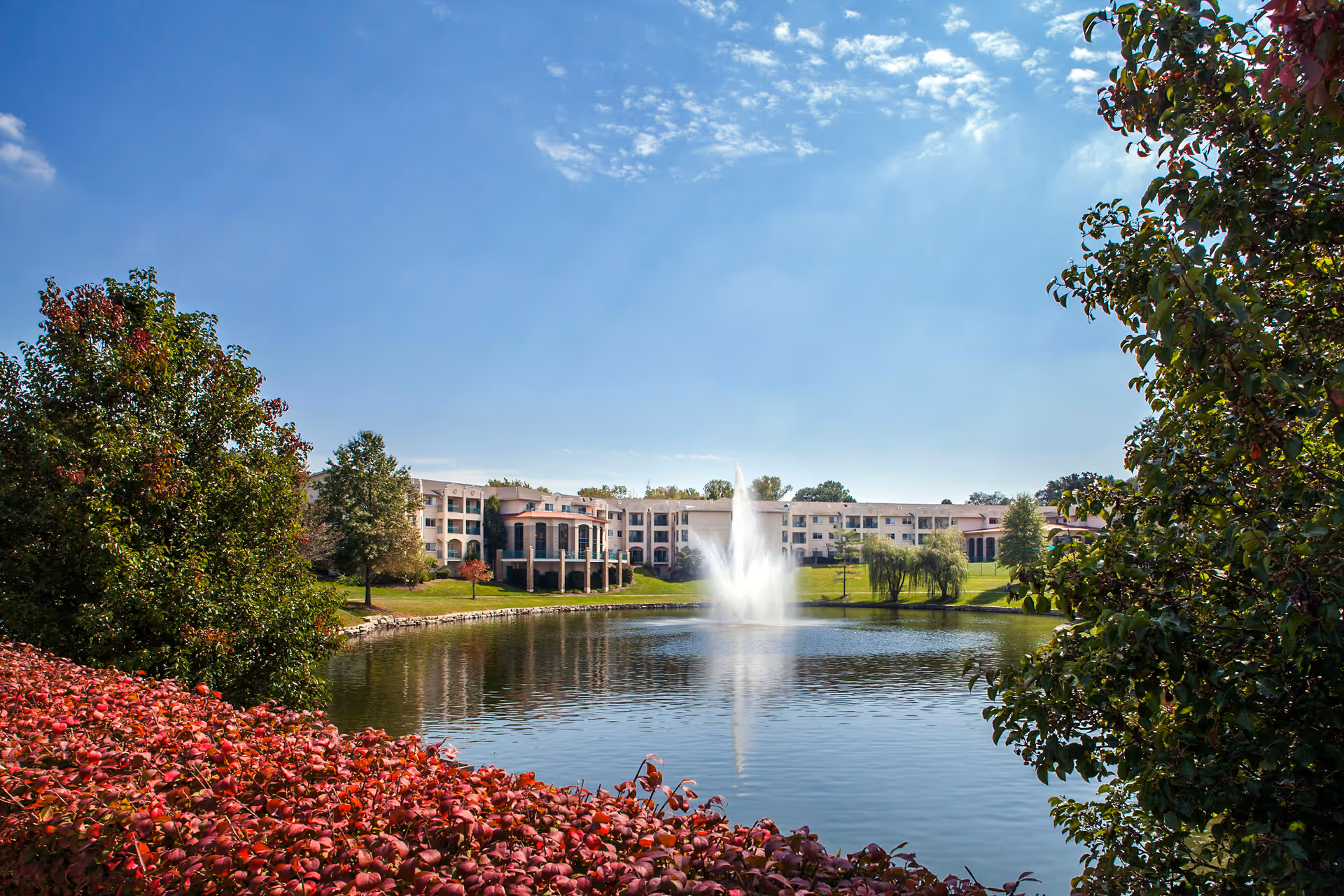 View of a senior living facility named Garden Villas North with a large pond and a water fountain in the center, surrounded by green trees and bushes under a clear blue sky.