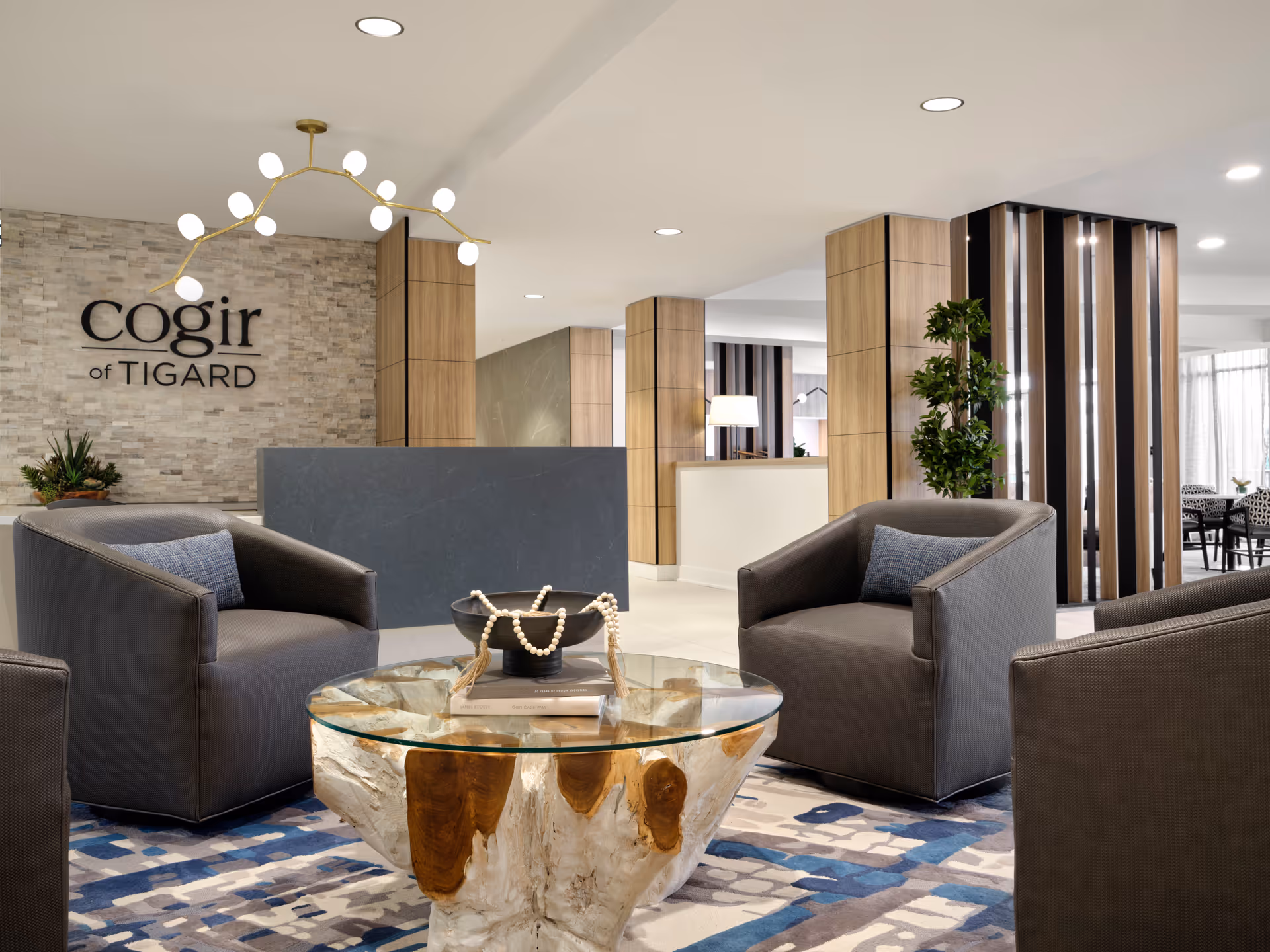 Modern senior living facility lobby with a stone accent wall displaying the sign 'cogir of TIGARD'. The seating area includes three gray armchairs with blue cushions arranged around a glass-top coffee table with a natural wood base. The space features wooden columns, a contemporary chandelier, a patterned blue and gray rug, and some greenery, creating a welcoming and stylish atmosphere.