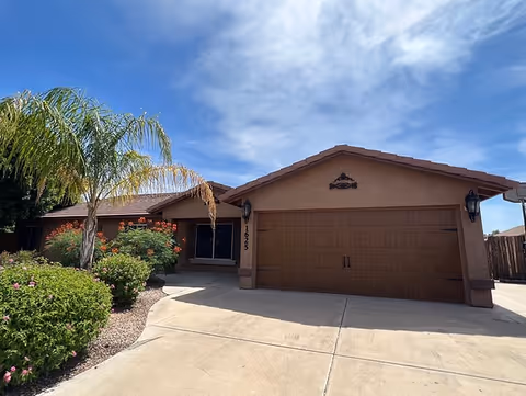 Front exterior view of a single-story residential building with a brown garage door, a driveway, and landscaped greenery including palm trees and bushes under a partly cloudy sky.