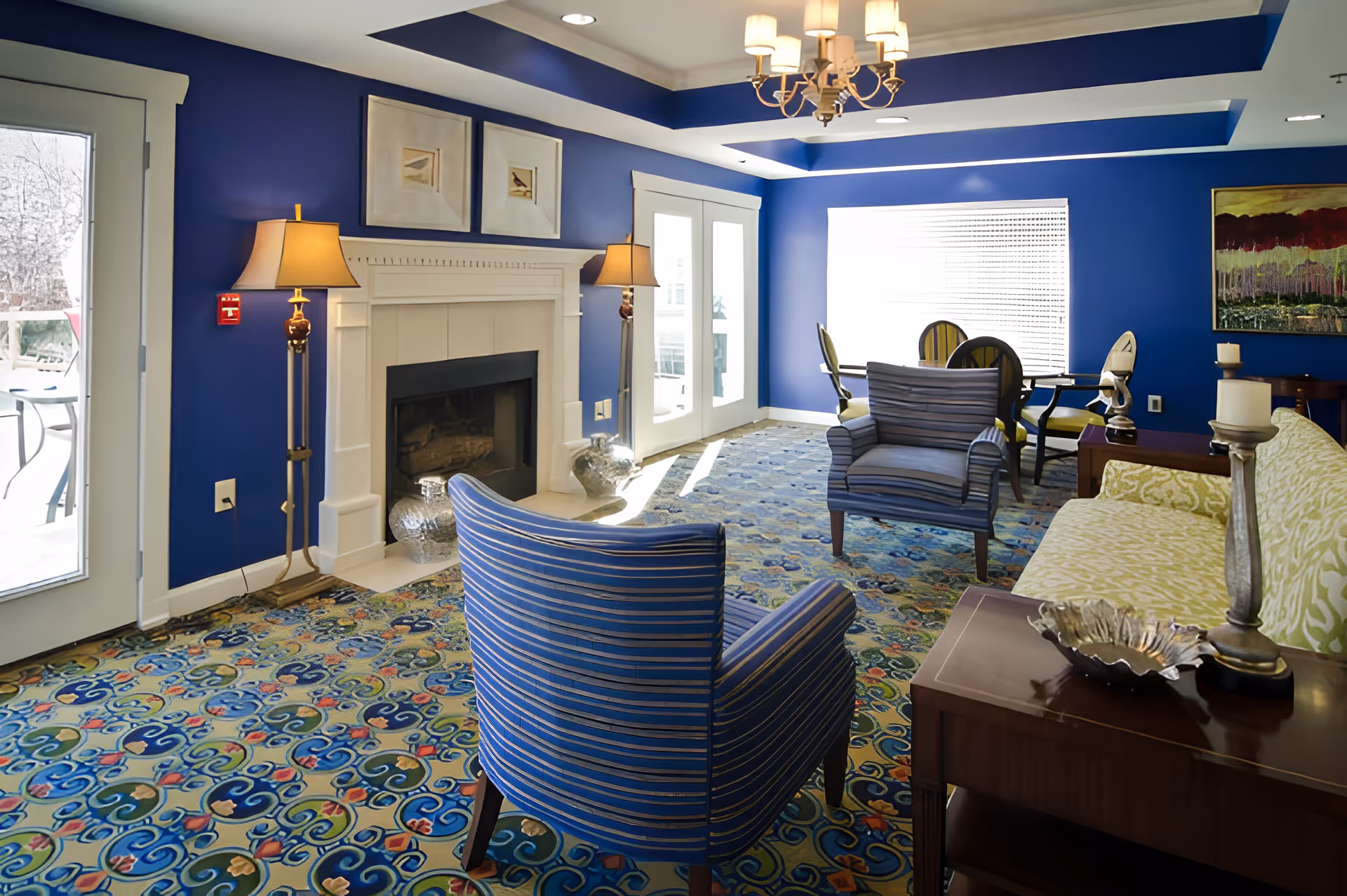 A cozy living room with bright blue walls and a patterned carpet. The room features a white fireplace with two lamps on either side, two blue striped armchairs, a green patterned sofa, a wooden side table with decorative items, and a dining table with chairs near a large window with blinds. A chandelier hangs from the ceiling, and there are two framed pictures above the fireplace.
