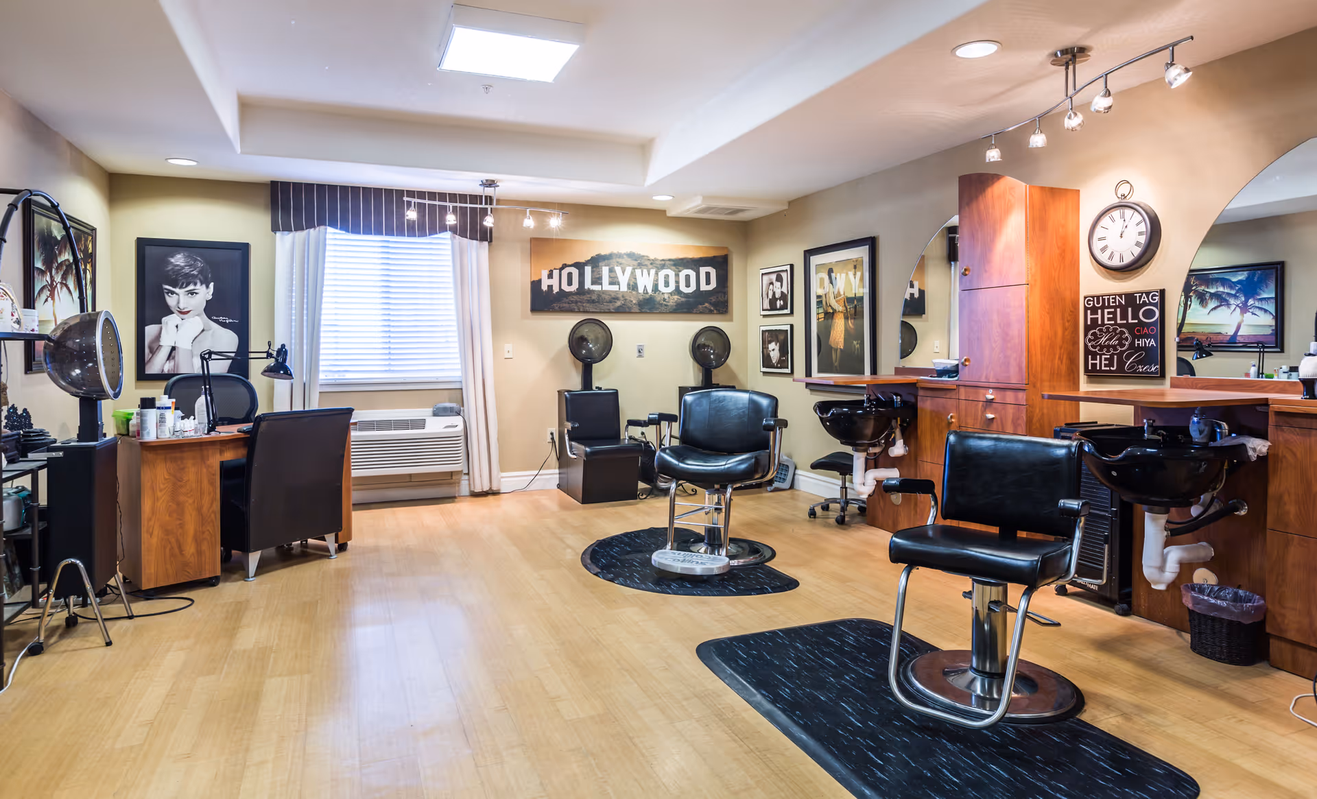 Interior of a hair salon with wooden flooring, two black salon chairs on black mats, hair washing stations with sinks, a desk with a chair and hair styling tools, framed black and white photos on the walls, a window with white curtains, and a sign that reads 'HOLLYWOOD'.
